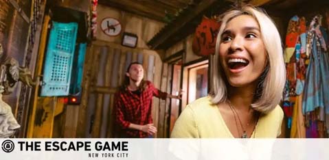 Image shows two individuals in a room with rustic decor typical of an escape room activity. A woman in the foreground is smiling broadly with excitement, while a man in the background appears to be searching for clues. The logo at the bottom indicates it's 'The Escape Game' in New York City.