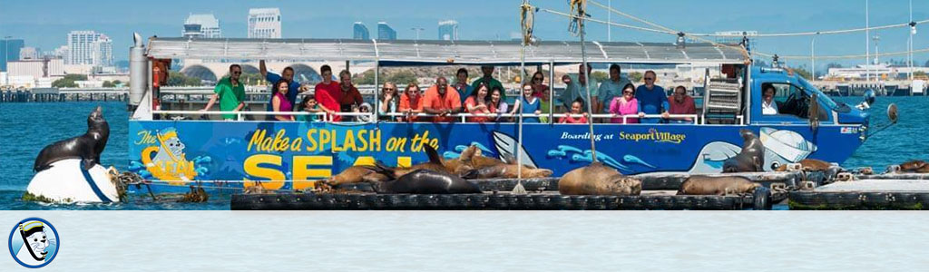 Tour boat with passengers near sea lions on a floating dock, city skyline in the background.