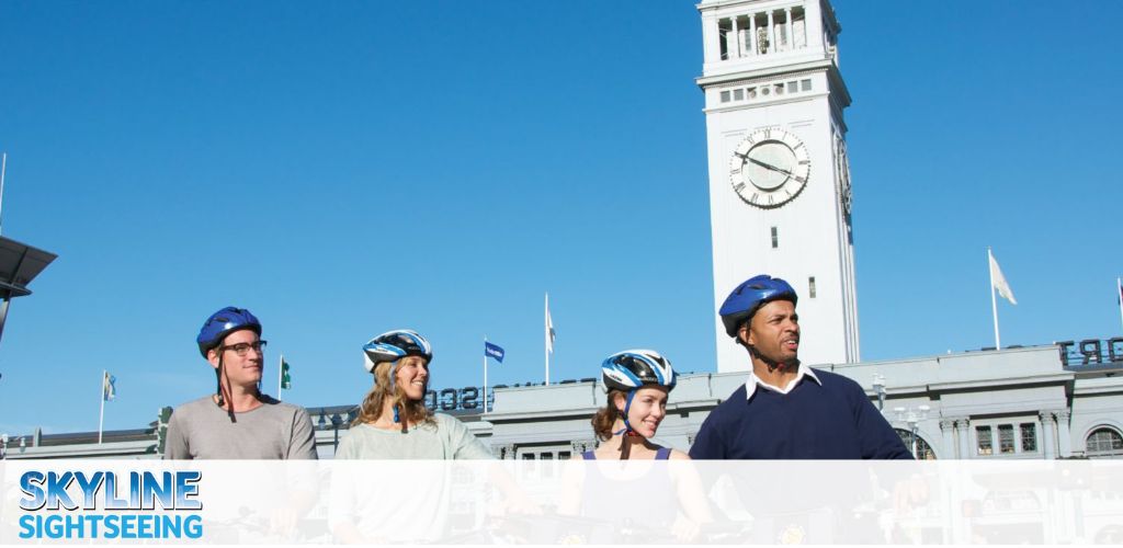This image displays a clear, sunny day with a bright blue sky as the backdrop. Four individuals, each wearing blue safety helmets, stand side by side in the foreground with a pleasant, engaged demeanor. They appear to be on a sightseeing adventure, possibly a bike tour, given their attire. In the center background, an iconic white clock tower with a classic architectural style stands tall. The time on the clock is visible, suggesting that it's midday. The words "Skyline Sightseeing" are superimposed on the image, indicating that this scene is likely part of a sightseeing tour promotion. The structures suggest an urban area, and flags can be seen fluttering atop the building behind the clock tower. 

At GreatWorkPerks.com, we are committed to bringing you the joy of adventure with the added bonus of savings; shop with us to find the lowest prices on tickets for your next sightseeing experience!