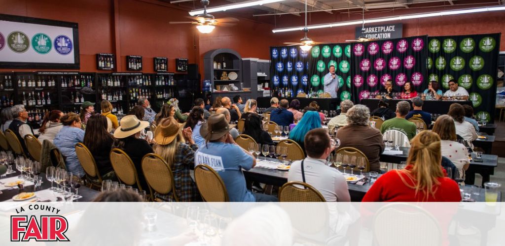 This image depicts an indoor event at what appears to be the LA County Fair. A diverse group of attendees, many wearing hats, are seated at long tables facing a panel at the front of the room. Each table has several rows of empty wine glasses set up in preparation for what may be a wine tasting event or educational workshop, given the context of the setup. The panel consists of five individuals, standing behind a table decorated with a row of barrels and an assortment of bottles on shelves behind them. The entire scene is illuminated by overhead lighting that gives off a warm ambiance. A large banner above the panelists reads "Marketplace Education," suggesting an informative aspect to this gathering. Various award ribbons with "LA County Fair" embellish the backdrop, adding to the festive and competitive atmosphere typically associated with county fairs.

At FunEx.com, we believe in enriching your experiences with valuable learning opportunities and memorable moments. Sign up with us for the lowest prices on tickets and enjoy exclusive savings on your next fair adventure!