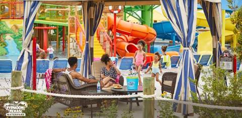 Visitors relax in shaded cabana seating at Typhoon Texas Waterpark, with colorful slides and playful water structures in the background amidst a lively, sunny setting.