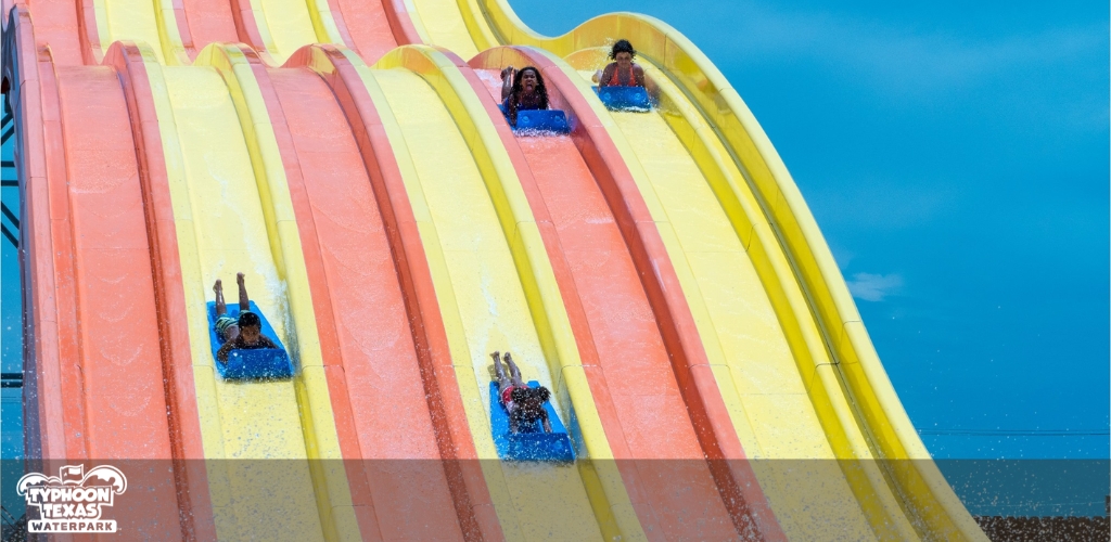 Visitors are enjoying a multi-lane water slide at Typhoon Texas Waterpark. There are four brightly colored slides, side by side, in hues of orange, yellow, and pink. People in blue rafts are sliding down, with water splashing around as they race toward the bottom under a clear blue sky.