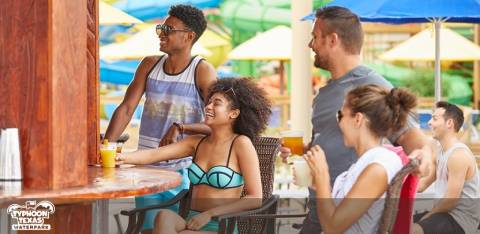 Group of diverse people enjoying a sunny day at an outdoor bar. Men and women are casually dressed, suitable for a waterpark, some sitting and standing, with drinks in hand, umbrellas in the background suggesting a relaxed, vacation atmosphere.