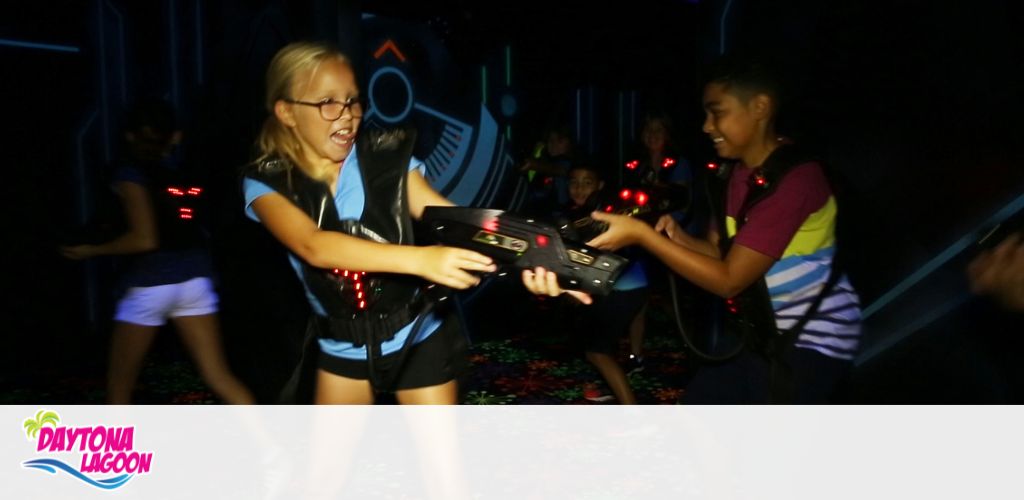 Two kids in vests play laser tag, one smiling, with dim neon lighting.