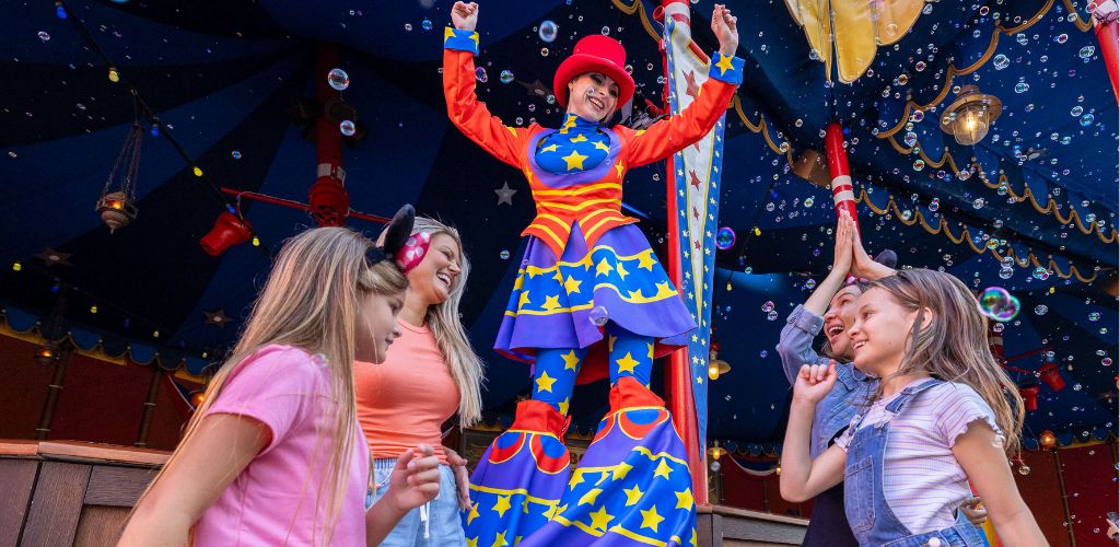 A performer dressed as a colorful clown stands on stilts entertaining children inside a decorated circus tent with bubbles floating around.