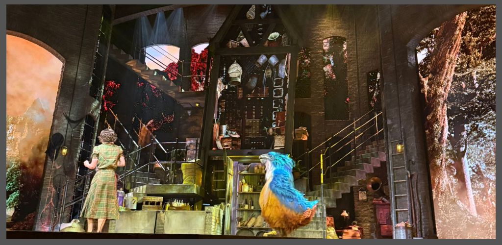 A colorful stage with a person in a plaid dress and a large puppet bird in front of books and art supplies against a forest backdrop.