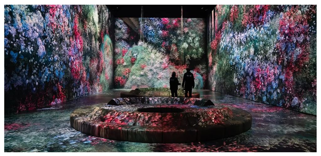 Two people stand inside a dark room with large colorful floral projections on the walls and floor, creating an immersive and vibrant visual experience.