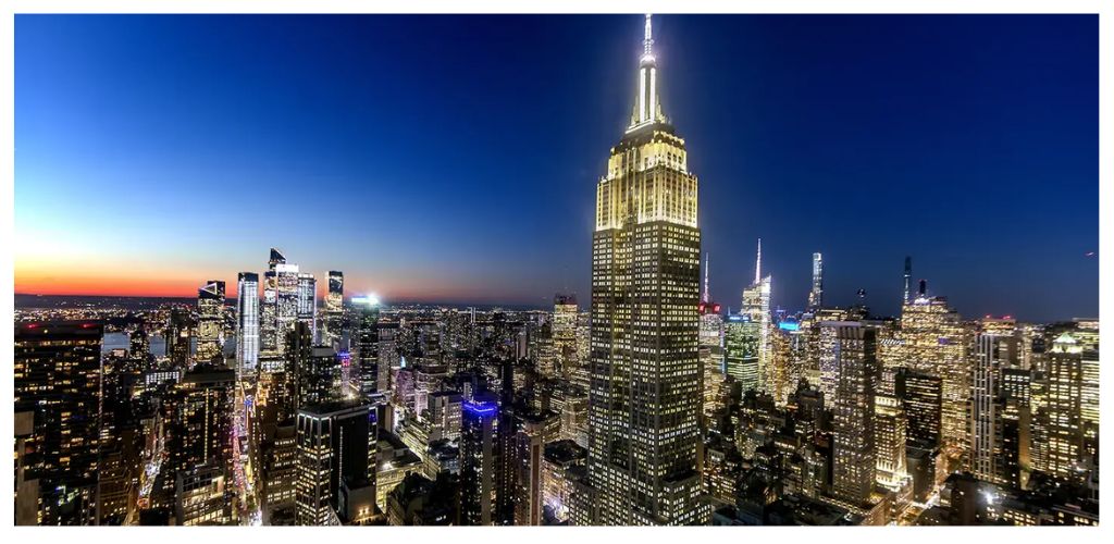 A nighttime cityscape featuring the illuminated Empire State Building surrounded by tall skyscrapers in New York City