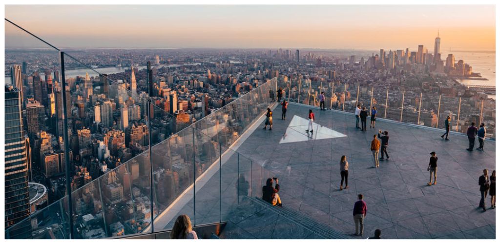 People viewing cityscape from rooftop observation deck with glass barriers, overlooking tall buildings and skyline during sunset