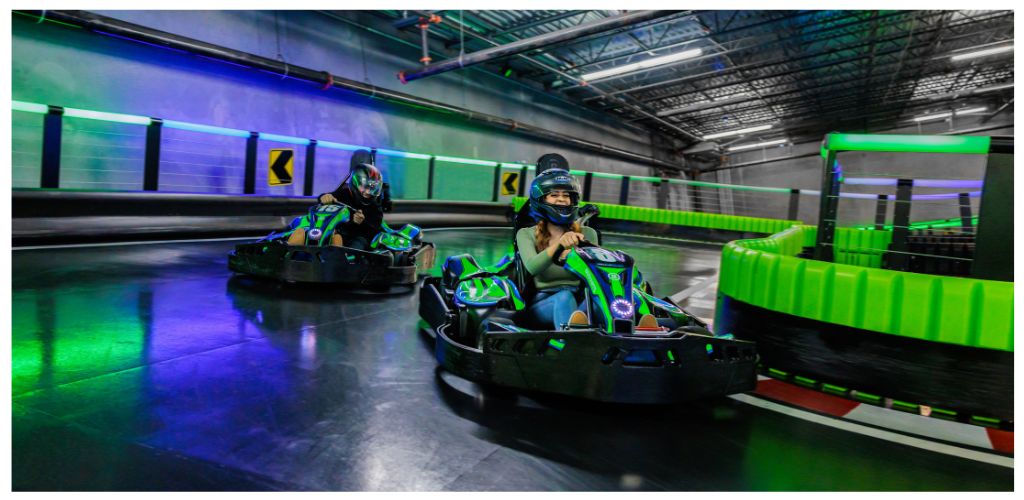 Two children wearing helmets and face masks ride go-karts on an indoor track with colorful neon lighting and safety barriers.