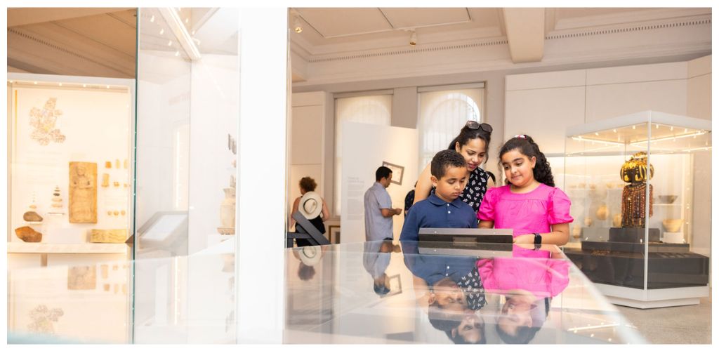 A woman and two children look at exhibits on a tablet in a museum with display cases of artifacts and artwork in the background.