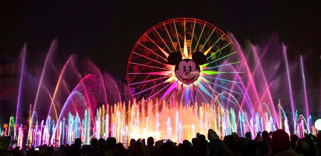 Nighttime image of a fireworks display with colorful jets, featuring a large Ferris wheel with Mickey Mouse’s face illuminated in the center, and a crowd watching below.
