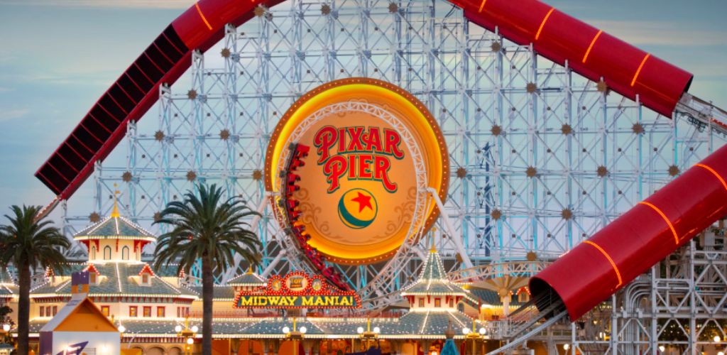 Pixar Pier at Disneyland featuring a large, lit-up sign, red roller coaster rides, and palm trees against the evening sky.