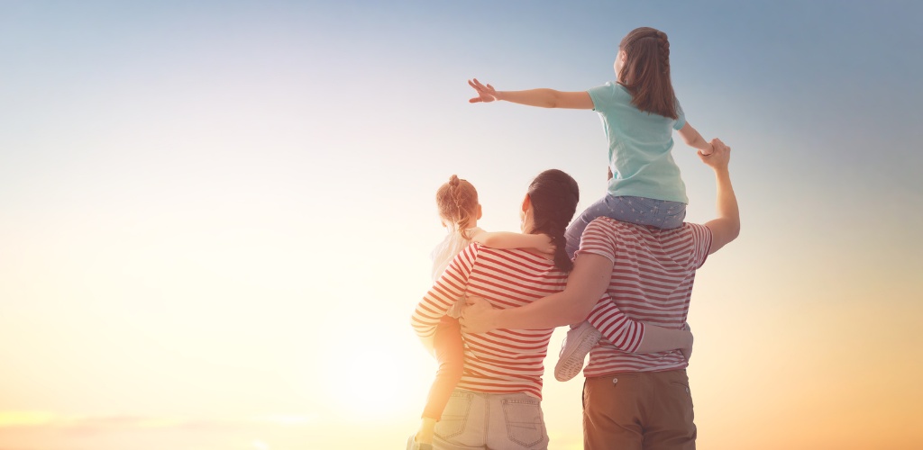 A family enjoying a sunset outdoors, with a child sitting on a man's shoulders and another child embracing a woman.