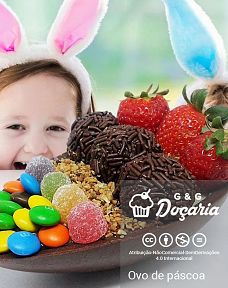 Duas crianças com orelhas de coelhinho apoiadas sobre uma mesa observam ansiosas um ovo de Páscoa de colher feito de chocolate com morangos, brigadeiros, castanhas trituradas, balas de goma e M&M's, que está sobre a mesa