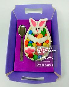 Ovo de Páscoa de colher feito de chocolate branco, decorado com um coelhinho segurando uma cenourinha, ambos feitos em pasta americana, mergulhado sobre o recheio coberto por M&M's, dentro de uma caixinha lilás com uma colherzinha metalizada