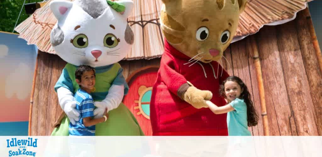 Two children stand smiling with life-sized cartoon cat characters in colorful costumes, positioned in front of a rustic, wooden backdrop at Idlewild and SoakZone. The boy, in a blue striped shirt, embraces a white and gray cat character, while the girl, in a turquoise shirt, holds hands with a brown cat character in a red outfit. The scene conveys joy and playfulness.