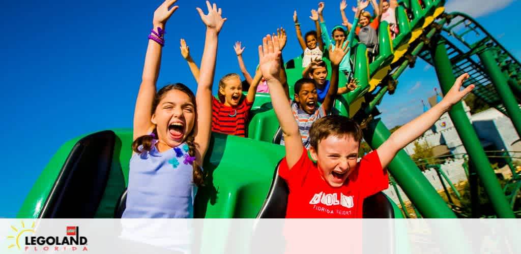 Excited children are raising their hands in joy on a vibrant green LEGOLAND Florida roller coaster under a clear blue sky, expressing the thrill of the ride. The LEGOLAND Florida logo is visible in the corner.