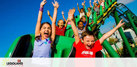 Children on a Legoland roller coaster are shown in the image. Their arms are raised and faces express excitement. The background has bright blue sky and parts of the green track. The Legoland logo is visible in the corner.
