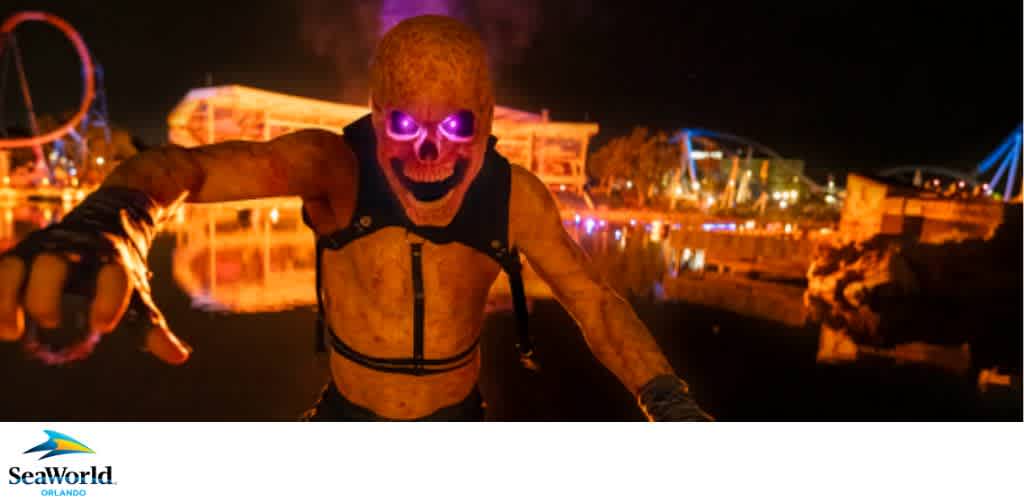 A person in a Halloween costume with a glowing skull mask stands in front of illuminated theme park attractions at night.