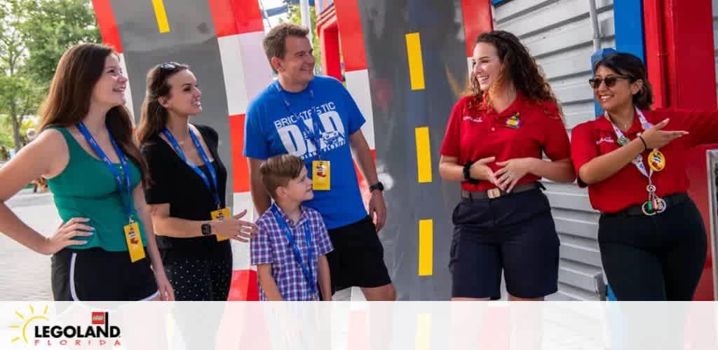 A joyful group interacts outside at LEGOLAND Florida. Two LEGOLAND employees in red uniforms engage with three guests: a man in blue, a woman in teal, and a boy. All exhibit smiles and attentive body language, with the LEGO themed backdrop enhancing the playful atmosphere.