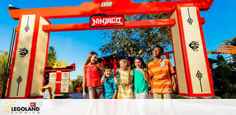 A family of five smiles in front of a large red and yellow Legoland Ninjago themed entryway adorned with ninja symbols under a bright blue sky. A logo on the bottom indicates the location as Legoland.