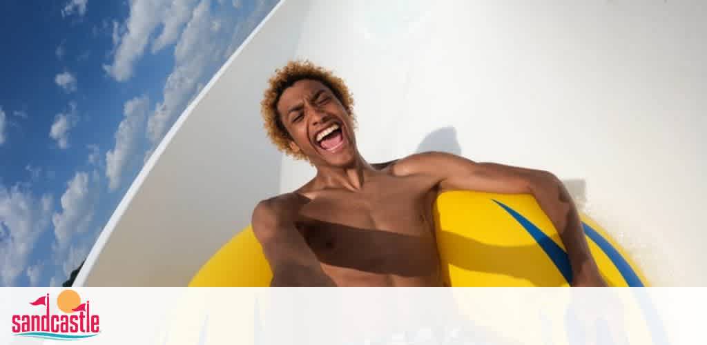 Image depicts a person with a joyful expression riding a waterslide. The person is holding onto a yellow raft with blue stripes. The backdrop has a clear blue sky with some clouds. The Sandcastle logo is visible in the lower left corner.