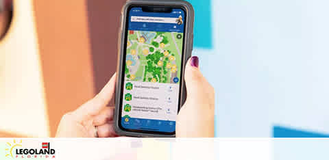 A person's hands hold a smartphone displaying a map application, presumably of a theme park, with various points of interest marked. The phone's case is blue and a part of the person's purple manicure is visible. The logo at the bottom suggests the map is of LEGOLAND. The background is blurred, emphasizing the phone.
