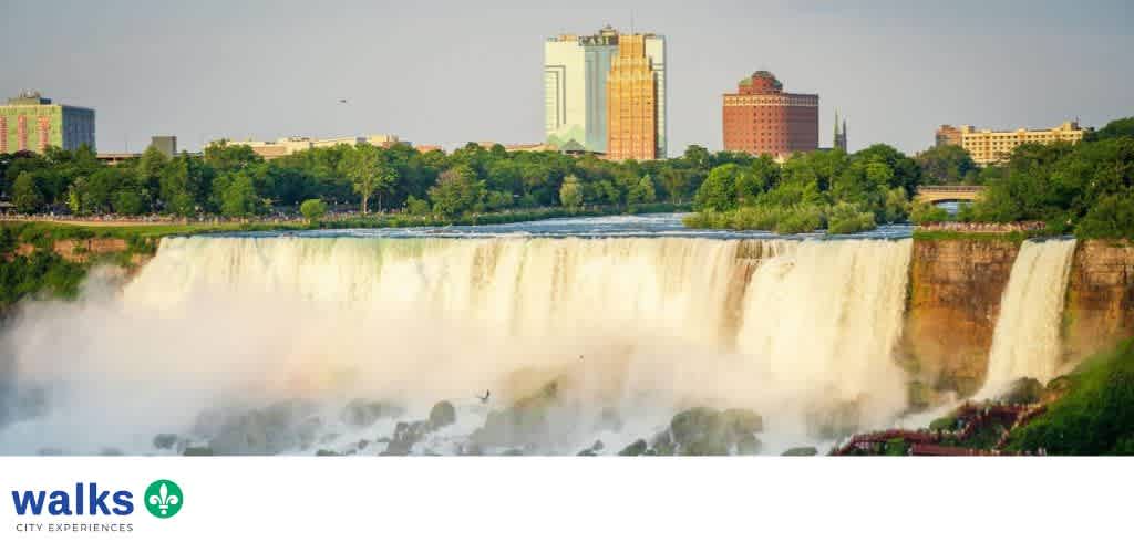 A wide view of a large waterfall with mist rising, city buildings in the background, and lush green trees along the riverbank