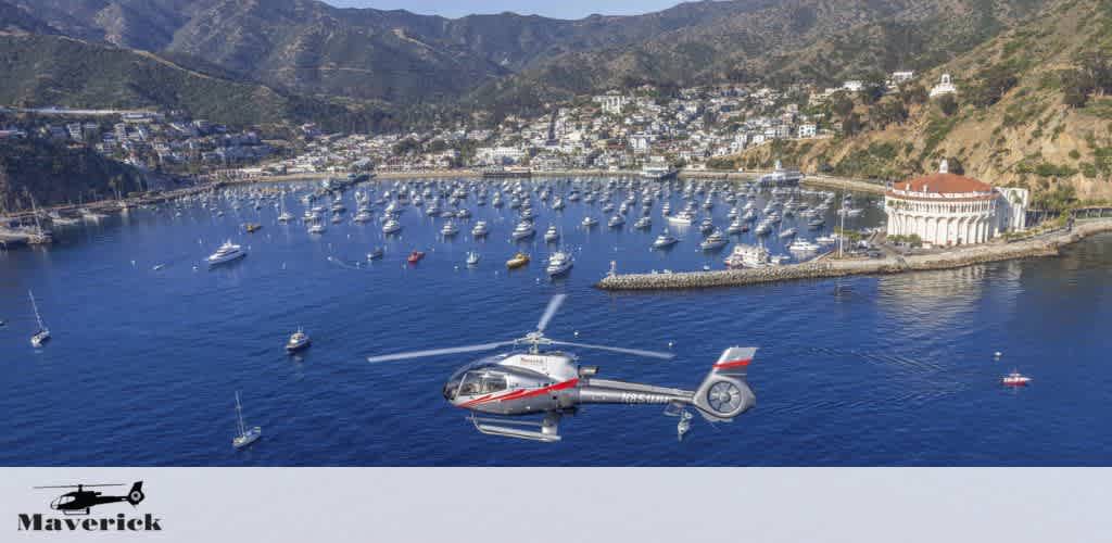 Aerial view of a sunlit harbor with a dense cluster of boats. A helicopter branded Maverick flies in the foreground over the water. The backdrop features a hilly landscape with buildings. A white, columned structure is seen on the right.