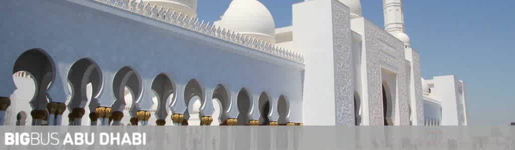 Panoramic view of an iconic white mosque with domes and arches under a clear blue sky. Text overlay at the bottom reads: BIGBUS ABU DHABI.