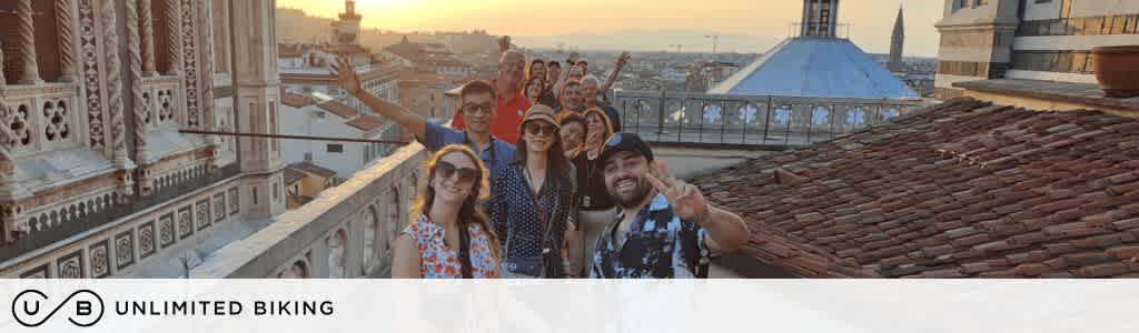 A group of smiling people stand on a rooftop terrace overlooking a cityscape at sunset. They are posing for a photo, with some making peace signs. The background features historic buildings and a blue dome. The image promotes Unlimited Biking.