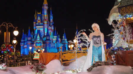 Queen Elsa from Frozen standing in a snowy scene in front of Cinderella Castle illuminated with winter lights during a Disneyland holiday celebration.