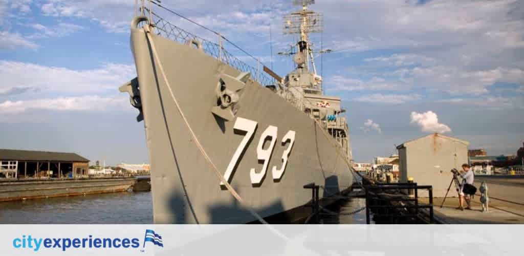 Image shows a large navy ship with the number 793 displayed on its hull, docked at a pier under a clear sky. Two individuals appear on the right, one seemingly adjusting a tripod. The logo for City Experiences is visible on the top right corner.