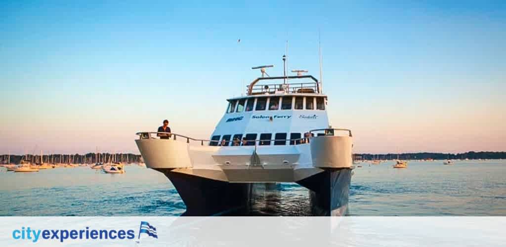Image shows a large white ferry with 'City Experiences' branding on its side, anchored near a harbor at sunset. A person stands on the front deck looking out over the water where multiple boats are visible in the distance, under a soft blue sky.