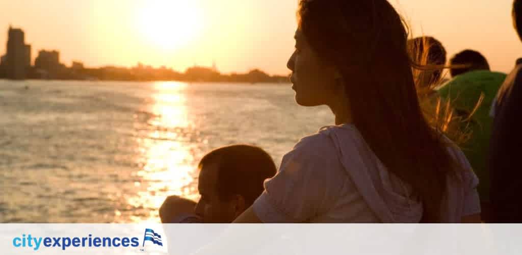 Silhouetted figures against a sunset over water with a cityscape backdrop. A woman looks forward while holding a child, her hair lit by the fading sun. The City Experiences logo is on the bottom right.