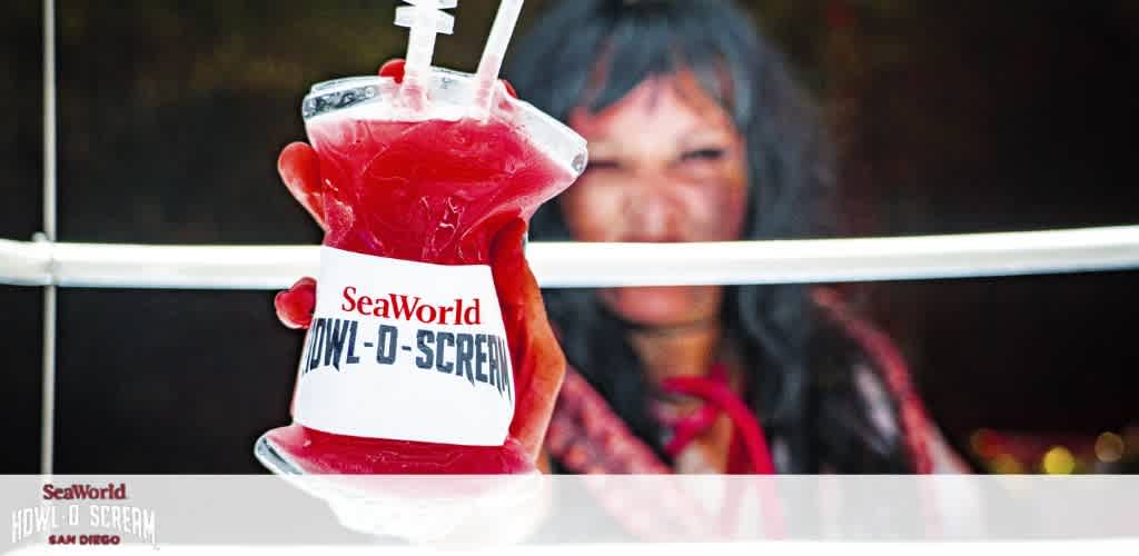 Image displays a hand holding a clear IV bag filled with a red beverage labeled 'SeaWorld HOWL-O-SCREAM'. A blurred figure in what seems like Halloween attire is in the background. Logo and event details are visible on the bottom of the image.