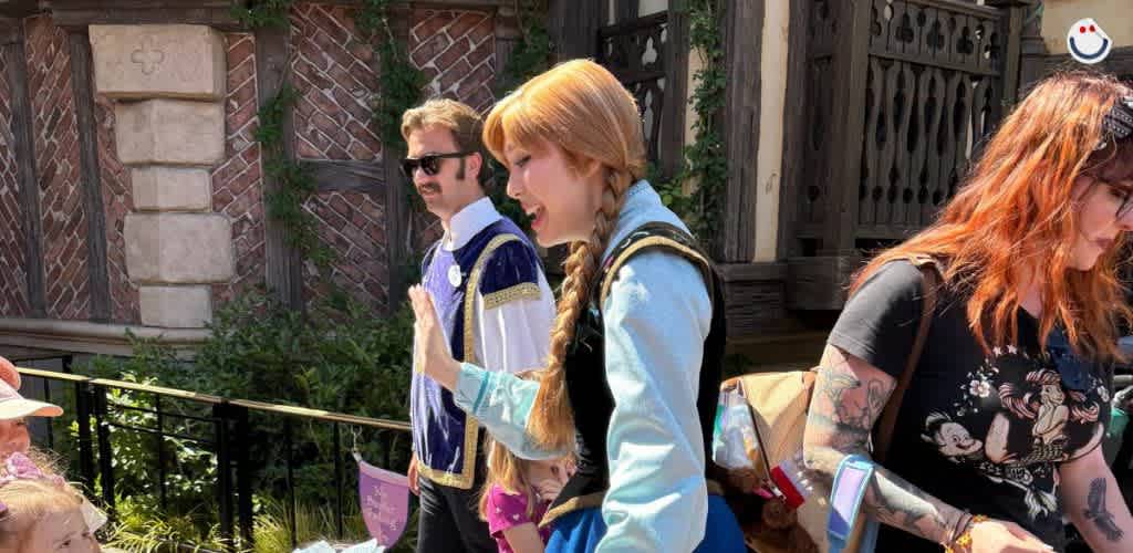 A costumed character in a blue dress interacts with children outdoors, accompanied by a person in a medieval-style outfit and a woman with tattoos. They stand in front of a rustic building with ivy-covered walls. The atmosphere is lively and welcoming, perfect for family enjoyment.