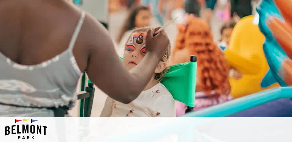 Image Description: This is a vibrant photo taken at an event at Belmont Park. In the foreground, a child is sitting patiently, slightly turned away from the camera, while an adult, whose back is facing us, applies face paint with care and precision. The child appears to be enjoying the experience, with a beautiful design that includes red and white colors and a spiraling pattern on the forehead. The background is softly out of focus, suggesting a lively event atmosphere with bright colors, possibly balloons or other festive decorations, lending to the overall feeling of a family-friendly environment. The logo of Belmont Park is visible in the corner of the image.

Don't miss out on memorable experiences at Belmont Park; visit GreatWorkPerks.com to secure your tickets at the lowest prices, offering you great savings on your next family outing!