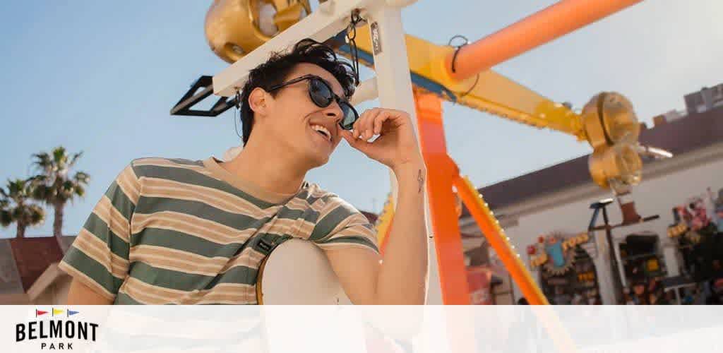 This image shows a person with short dark hair, wearing sunglasses and a green and gray striped shirt, smiling and enjoying themselves at Belmont Park. They appear to be sitting in an open-air ride compartment with a safety harness, which is part of a larger amusement park ride characterized by bright orange and yellow colors. The ride structure frames the photo to the left and upper part of the image. In the background, palm trees and clear blue skies suggest a sunny day, perfect for outdoor activities. The Belmont Park logo is visible in the bottom left corner, indicating the location.

At GreatWorkPerks.com, we're committed to bringing you the thrill of amusement parks at the lowest prices—discover our exclusive discounts and savings on tickets today!