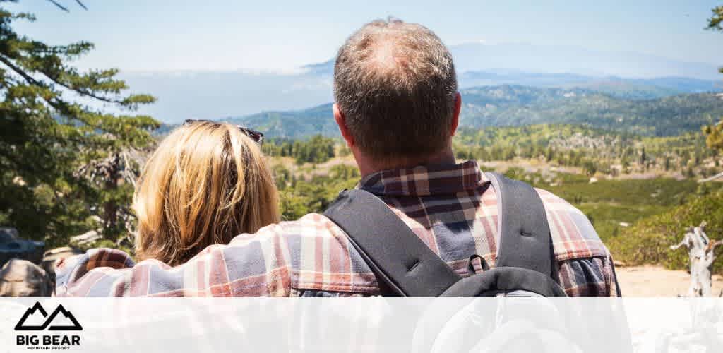 Two people in plaid shirts overlook a scenic mountain landscape.