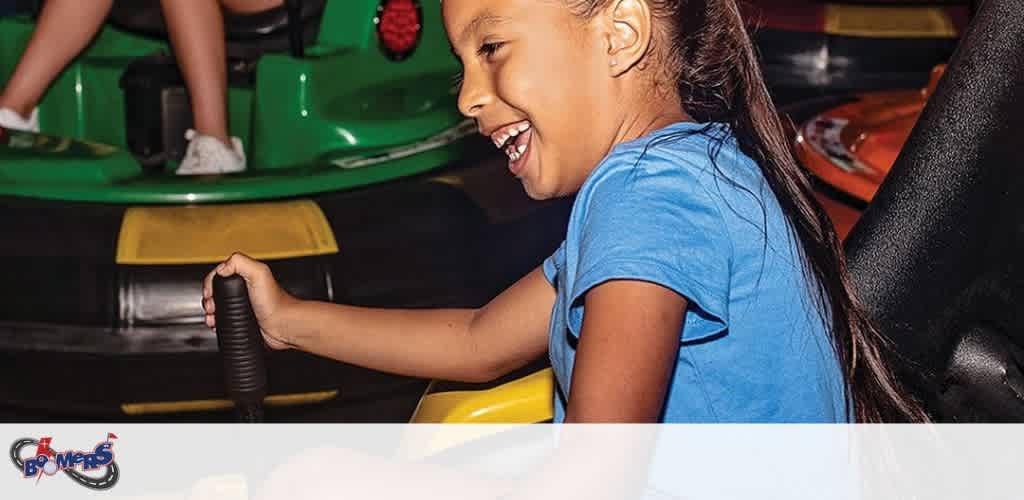 A joyful girl with long hair in a blue shirt enjoys driving a yellow bumper car at an indoor amusement park.