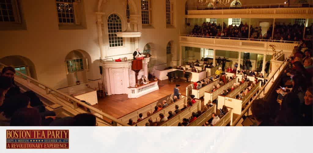 Image of an indoor historical reenactment at the Boston Tea Party Ships & Museum. Visitors occupy tiered wooden benches and balconies in a dimly lit, large hall. A central figure stands at a balcony pulpit, addressing the crowd. The atmosphere suggests an 18th-century setting.