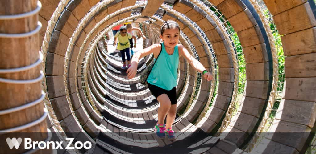 Image shows a child running through a curved, tunnel-like play structure made of wooden slats with a light-filled exit ahead. Another child is seen in the background. The structure is at the Bronx Zoo, as indicated by their logo at the bottom right of the image.