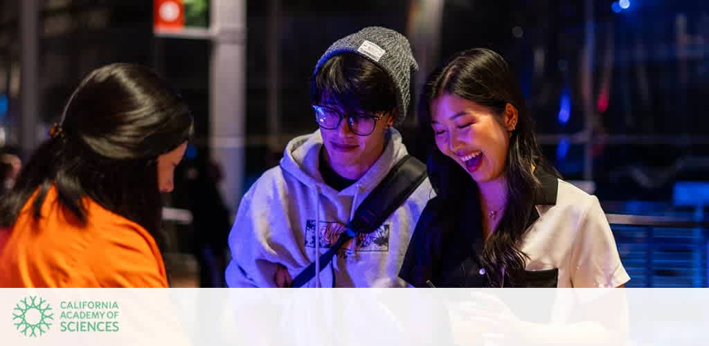 Three people smiling and looking down at an object, in an indoor setting with dim lighting. The California Academy of Sciences logo is visible in the bottom left corner.