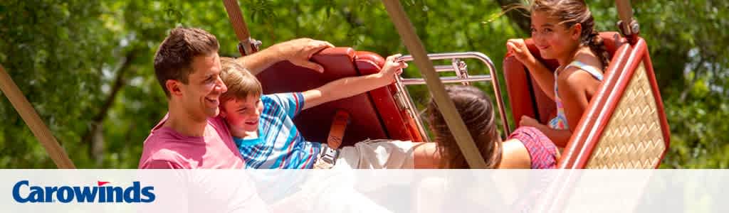 Image shows a man and two children smiling while riding a swing ride at Carowinds amusement park. Trees with green foliage are visible in the background, indicating a sunny day. The Carowinds logo is featured in the lower right corner.