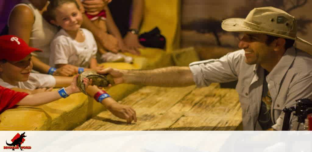 Guide presenting a small reptile to a smiling child in a hat among an engaged audience.