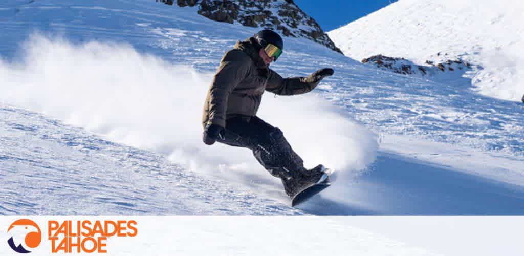 A snowboarder in winter gear descends a snowy slope with mountains in the background under a clear blue sky. Logo in corner reads