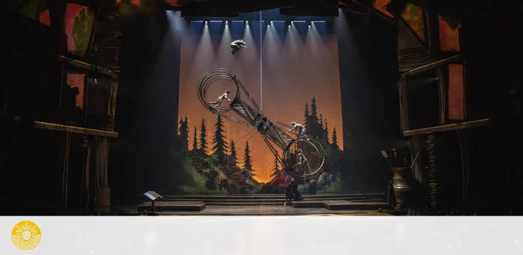 Theatrical stage with acrobats performing on a large double wheel in front of a backdrop of silhouetted trees.
