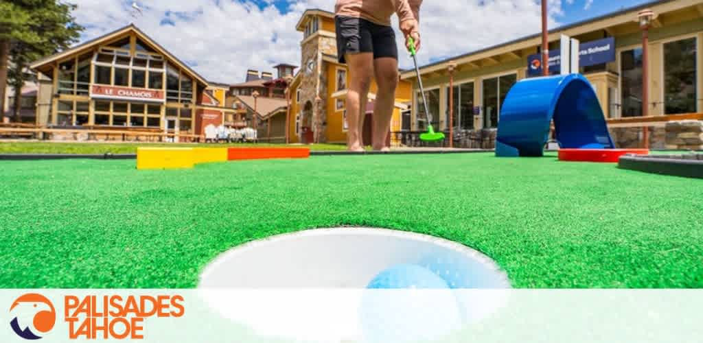 Mini-golf course with a focus on a blue ball near a hole; person with a green putter in the background.